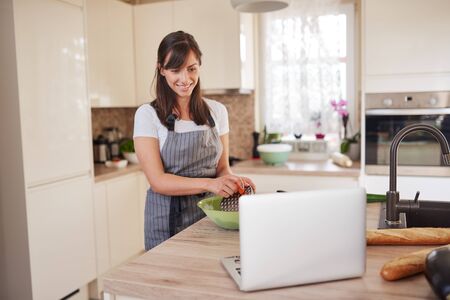 Young Caucasian brunette in apron grating carrots in bowl, preparing dinner and looking at lapotp while standing in kitchenの写真素材