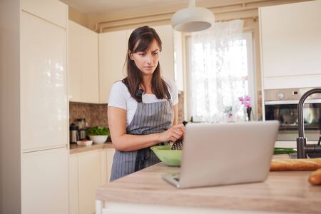 Young Caucasian brunette in apron grating carrots in bowl, preparing dinner and looking at lapotp while standing in kitchenの写真素材