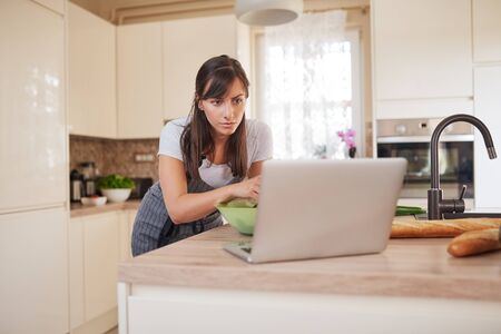 Pretty Caucasian brunette in apron leaning on kitchen counter and looking at laptop and following recipe for dinner.の写真素材
