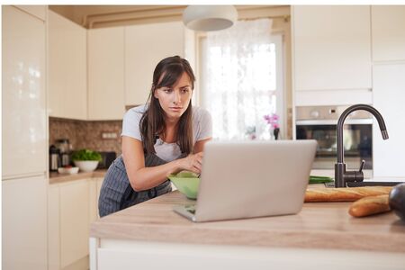 Pretty Caucasian brunette in apron leaning on kitchen counter and looking at laptop and following recipe for dinner.の写真素材