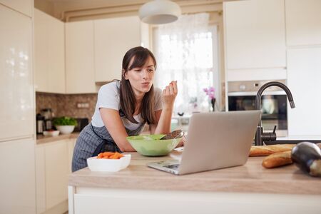 Gorgeous Caucasian brunette in apron leaning on kitchen counter, looking at laptop, looking for new recipe and eating carrot. On table is lots of vegetables.の写真素材