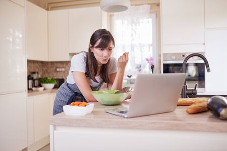 Gorgeous Caucasian brunette in apron leaning on kitchen counter, looking at laptop, looking for new recipe and eating carrot. On table is lots of vegetables.の写真素材