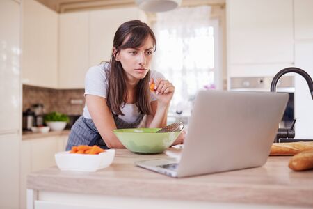 Pretty Caucasian brunette in apron leaning on kitchen counter and looking at laptop and following recipe for dinner. Woman holding carrot in hand.の写真素材