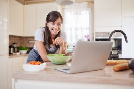 Pretty Caucasian brunette in apron leaning on kitchen counter and looking at laptop and following recipe for dinner. Woman holding carrot in hand.の写真素材