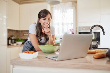 Pretty Caucasian brunette in apron leaning on kitchen counter and looking at laptop and following recipe for dinner. Woman holding carrot in hand.の写真素材