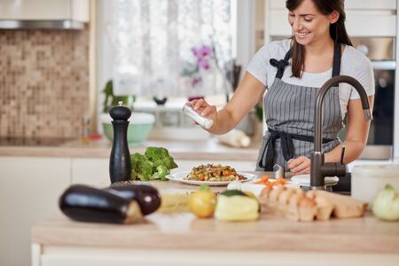 Cute Caucasian brunette in apron standing in kitchen and adding salt in fresh delicious pasta. Preparation of italian food concept.の写真素材