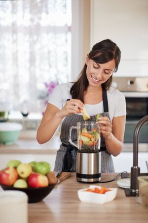 Beautiful Caucasian smiling woman in apron putting vegetables and fruits in blender while standing in kitchen.の写真素材