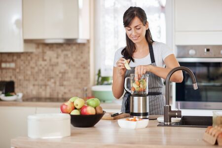 Beautiful Caucasian smiling woman in apron putting vegetables and fruits in blender while standing in kitchen.の写真素材