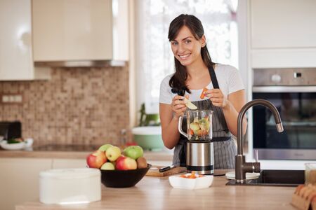 Beautiful Caucasian smiling woman in apron putting vegetables and fruits in blender while standing in kitchen.の写真素材
