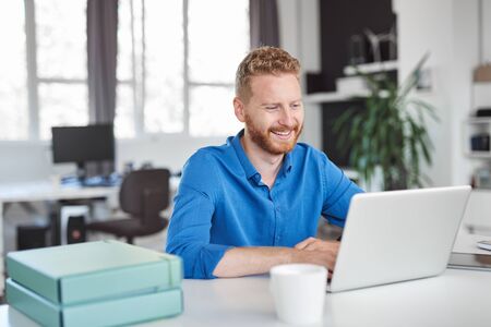Young smiling handsome Caucasian male employee sitting in office and using laptop. Start up business concept.の写真素材