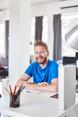 Cheerful Caucasain ginger employee sitting in his office and writing his tasks in notebook.の写真素材