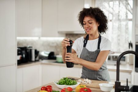 Attractive mixed race woman in gray apron addinf pepper in bowl with rocket and mushrooms. On...