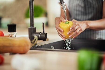 Close up of mixed race woman in apron standing in kitchen ad washing yellow paprika in sink.の写真素材