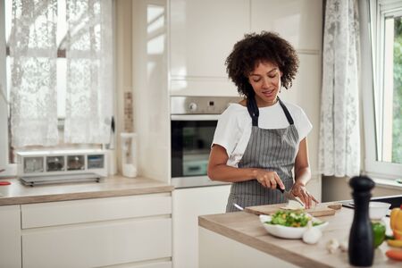 Gorgeous mixed race housewife in apron standing in kitchen and chopping mushrooms. On kitchen counter are all sorts of vegetables.の写真素材