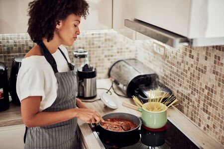 Mixed race woman in apron standing next to stove in domestic kitchen and making pasta and sauce.の写真素材