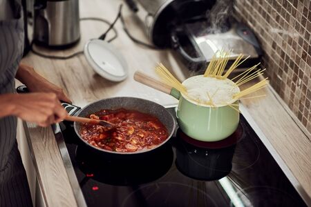 Mixed race woman in apron standing next to stove and stirring tomato sauce. On stove are saucepan and pot with spaghetti.の写真素材