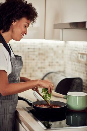 Mixed race woman in apron standing next to stove, stirring tomato sauce and adding rocket. On stove is pot with boiling pasta.の写真素材
