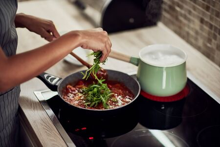 Mixed race woman in apron standing next to stove, stirring tomato sauce and adding rocket. On stove is pot with boiling pasta.の写真素材
