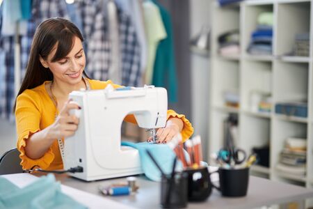 Smiling charming caucasian fashion designer sitting in her studio and sewing beautiful evening...