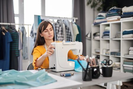 Focused charming caucasian fashion designer sitting in her studio and sewing beautiful evening dress.の写真素材