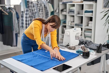 Focused attractive seamstress cutting textiles for beautiful night dress while standing in her...