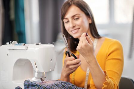 Attractive smiling caucasian brunette sitting in tailoring studio and cutting thread.の写真素材
