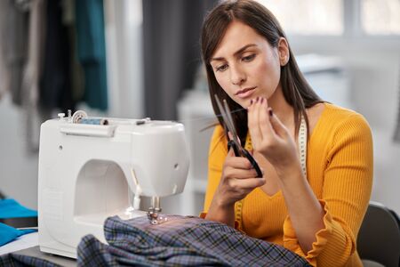 Attractive smiling caucasian brunette sitting in tailoring studio and cutting thread.の写真素材