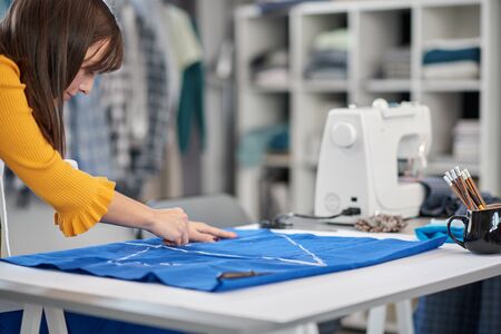 Creative caucasian fashion designer standing in her studio and drawing scheme on blue linen for a...