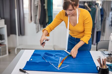 Dedicated seamstress standing next to table and cutting textiles for elegant dress.