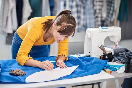 Creative caucasian fashion designer standing in her studio and drawing scheme on blue linen for a beautiful evening dress.の写真素材