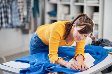 Creative caucasian fashion designer standing in her studio and drawing scheme on blue linen for a beautiful evening dress.の写真素材