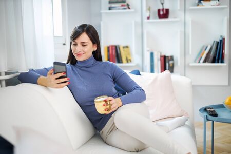 Young attractive smiling brunette sitting on couch in her living room, holding mug with coffee and hanging on social media.の写真素材