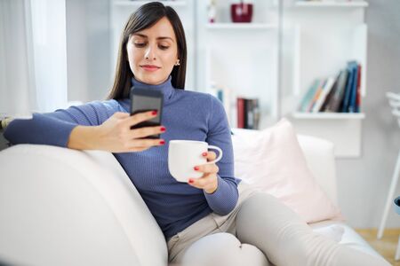 Young attractive smiling brunette sitting on couch in her living room, holding mug with coffee and hanging on social media.の写真素材