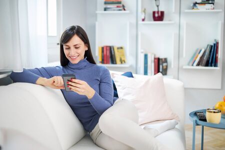 Young charming brunette sitting on couch in her beautiful home and typing message.の写真素材