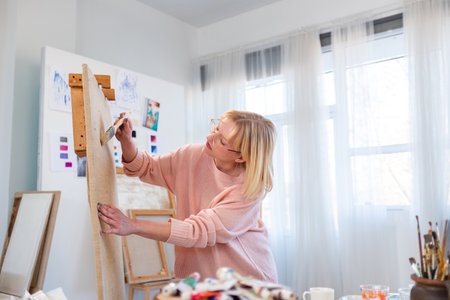 smiling senior woman painting picture on easel at home in sunny dayの写真素材
