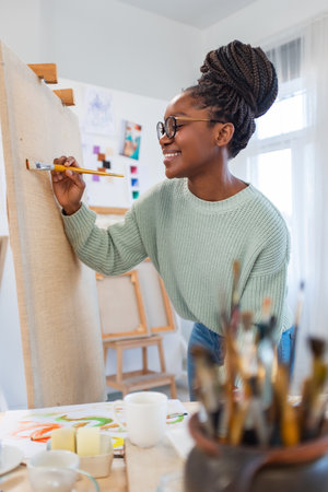 smiling african american woman painting on canvas in art studioの写真素材