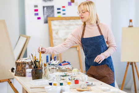 Mature female artist painting on canvas in art studio. She is standing at the table and holding paintbrushes.の写真素材