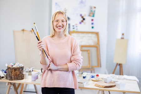 Portrait of smiling female artist holding paintbrushes while standing in studioの写真素材