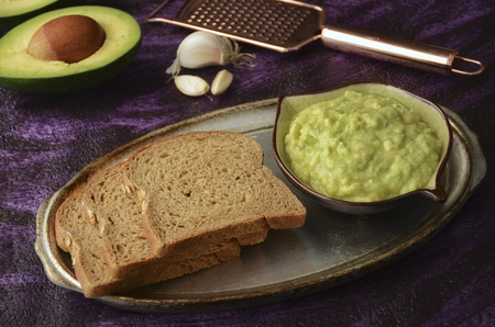 Guacamole and guacamole ingredients on purple background. Avocado, slice of bread, garlic and grater on plateの写真素材