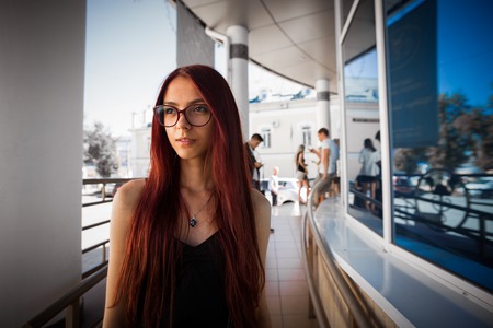 Urban city portrait of girl. Beautiful thinking girl with long red hair and glasses looking aside. Pretty girl in black dress on city, beautiful architecture background. Picture of pretty urban girlの写真素材