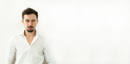 Young handsome man in white blouse stand on white background. Horizontal shot of serious stylish confident in white shirt man with beard. Man looking at camera while standing against white background. Man standing against white backgroundの写真素材
