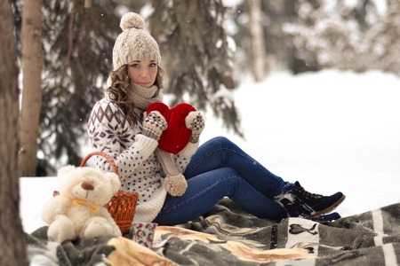 Attractive, beautiful, cute, pretty girl hold toy, girl with big, red heart in her hands, sit in warm plaid, has picnic in winter forest.Interesting, delightful, nice, attractive girl with big, read heart, toy in her hands in winter, snow, cold forest.の写真素材
