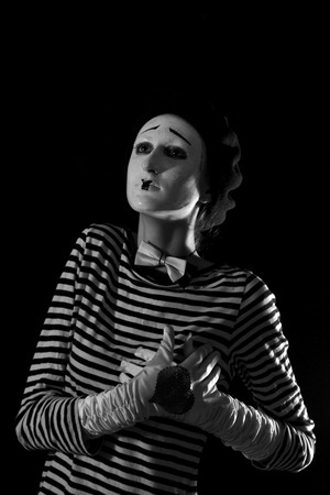 Emotional professional mime in stripped blacn and white shirt with black cap and white bow tie standing against black background in circusの写真素材