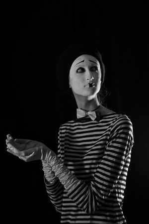 Emotional professional mime in stripped blacn and white shirt with black cap and white bow tie standing against black background in circusの写真素材