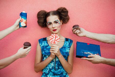 Beautiful fashionable girl with two hair bun a creative makeup holding a huge pink sweet lollipop in hands. Many hands on pink background round a girl holding cakes, juice, candies, sweets on backgroundの写真素材