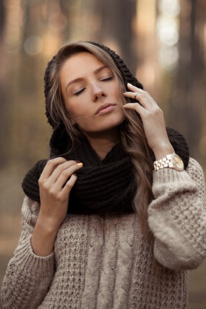 Fashionable, posing, stunning, attractive and very sensual, beautiful girl with closed elegant finders near the perfect face, pure skin.Inspiring, admiring, sensual, lovely, beautiful attractive girl in warm sweater and scarf in autum forestの写真素材