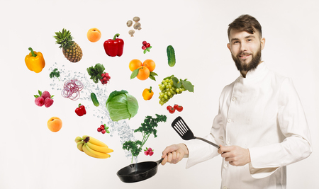Handsome professional chef in uniform uggling with vegetables and other food in the kitchen. Chef and flying vegetables and fruits osilated on white background. Fruits with vegetables and utensilsの写真素材