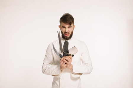 Studio shot of a happy bearded young chef holding sharp knives over white background. Chef with knife. Handsome angry serious cheef holding many sharp knives isolated against white background.の写真素材