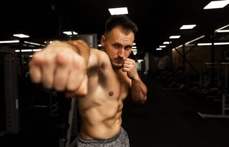 Young bearded handsome man with naked torso training alonein the dark sport gym. PUNCHING OT KICING...