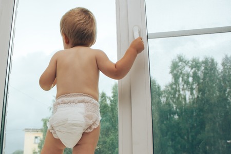 Cute baby boy is looking at camera while standing on the window sill at homeの写真素材